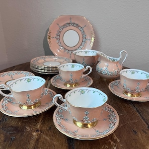 May include: A peach-coloured vintage tea set with a floral design. The set includes teacups, saucers, plates, a creamer, and a sugar bowl. Gold trim accents the edges of the pieces, creating an elegant aesthetic.