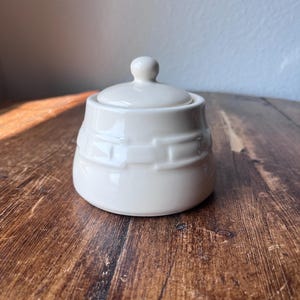 Puede incluir: Un azucarero de cerámica color crema con tapa a juego que presenta un pequeño pomo. El exterior del cuenco está decorado con un patrón de tejido de cesta en relieve que rodea la sección central del recipiente de forma redondeada.