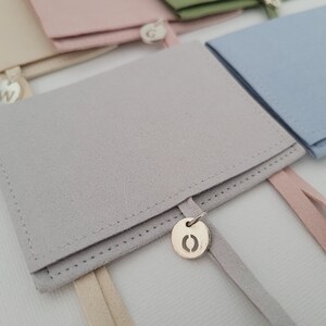 May include: A gray suede pouch with a cream-colored drawstring and a silver charm with the letter "O" on it. The pouch is folded in half and the charm is hanging from the bottom.