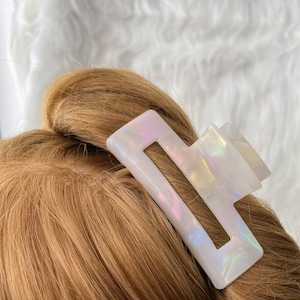 May include: A white rectangular hair claw clip with iridescent shimmer is placed in light brown hair. The clip has a geometric design with a rectangular shape and a cutout in the center. The background is a soft, white, fluffy texture.