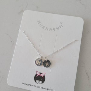 Pode incluir: Um colar de prata com dois pingentes redondos, cada um com uma letra, H e R, gravadas em ouro. O colar é apresentado em um cartão branco com o texto "MUSHROOM2" e "Instagram: mushroomsquared".