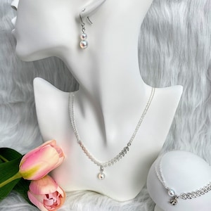 Op de afbeelding: Een witte parel ketting en armband set met een delicaat blad ontwerp. De ketting heeft een enkele parel hanger en de armband heeft een klein bedeltje.