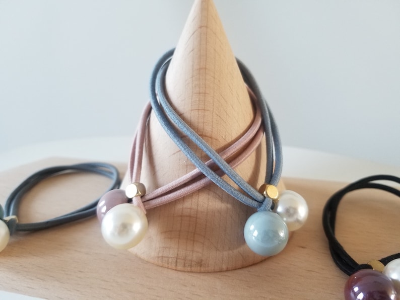 May include: Three hair ties with pearl and gold accents. The hair ties are in shades of blue, pink, and gray. The hair ties are displayed on a wooden cone.