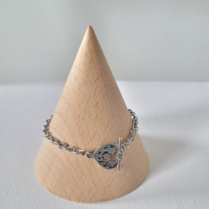 May include: A silver bracelet with a floral toggle clasp. The bracelet is displayed on a wooden cone.