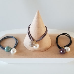 May include: Three hair ties with pearl and colored beads. The hair ties are displayed on a wooden stand. The beads are green, brown, and beige.