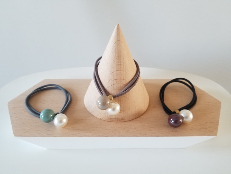 May include: Three hair ties with pearl and colored beads. The hair ties are displayed on a wooden stand. The beads are green, brown, and beige.