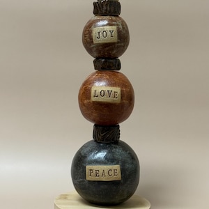 May include: A handcrafted ceramic sculpture featuring three stacked spheres. The spheres are brown, orange, and dark gray, with the words "JOY," "LOVE," and "PEACE" inscribed on them. The sculpture rests on a wooden base.