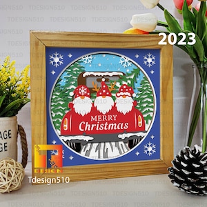 May include: A wooden framed picture with a blue background and a white circle containing three gnomes in a red car with the text "Merry Christmas". The year 2023 is written in the top right corner of the frame.