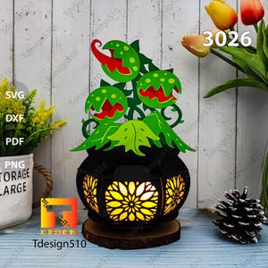 May include: A black and green plant lamp with a light inside. The plant has three mouths with red teeth. The lamp is on a wooden base. The number 3026 is visible in the image.