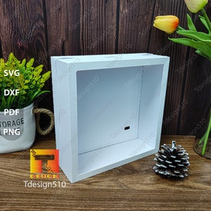 May include: A white, rectangular shadow box with a recessed interior. The box is made of a solid material and has a clean, modern design. A pinecone and a potted plant are nearby. The text "SVG, DXF, PDF, STORAGE, PNG" is visible.