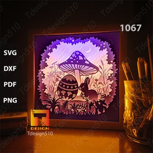 May include: A white frame with a purple, blue, and white paper cut design of a bunny rabbit, a mushroom, and an Easter egg. The design is illuminated from behind with a purple light.