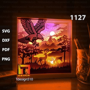 May include: A white frame containing a lit-up image of an eagle flying over a mountain range with deer in the foreground. The image is a paper cut-out design. The frame is sitting on a wooden surface. The text "Tdesign510" and "1127" are visible on the frame.