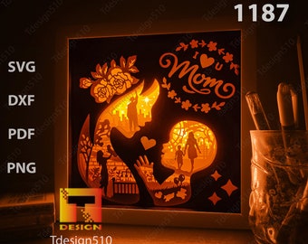May include: A white wooden frame with a cutout design of a mother and child. The design is illuminated from behind, creating a warm glow. The text "I Love You Mom" is featured in a decorative font.