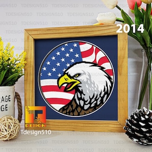 Puede incluir: Una impresión enmarcada de un águila calva americana con la cabeza girada hacia un lado, mirando hacia la izquierda. El águila está delante de una bandera americana circular con rayas rojas y blancas y estrellas azules. El marco es de madera y tiene un fondo azul oscuro. El año 2014 está impreso en la esquina superior derecha de la imagen.