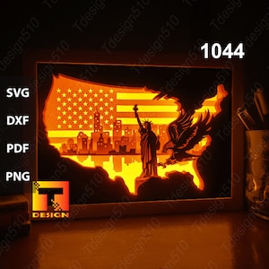 May include: A 3D papercraft light-up shadow box featuring a silhouette of the Statue of Liberty, an eagle, and the American flag. The shadow box is lit from behind, creating a warm glow.