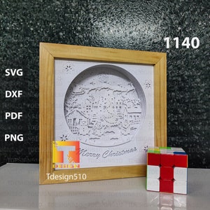 Puede incluir: Un dise&ntilde;o de papel recortado blanco de una escena de pueblo de invierno con el texto "Merry Christmas" en un marco de madera. El dise&ntilde;o est&aacute; en un c&iacute;rculo y tiene un efecto 3D. El marco est&aacute; sobre una superficie gris con un peque&ntilde;o cubo rojo y blanco.