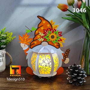 Peut inclure: Une lanterne de gnome en papier avec un corps blanc en forme de citrouille et un chapeau brun décoré de feuilles d'automne et de tournesols. La lanterne est éclairée de l'intérieur, projetant une lueur chaude. Le gnome tient une petite branche dans sa main.