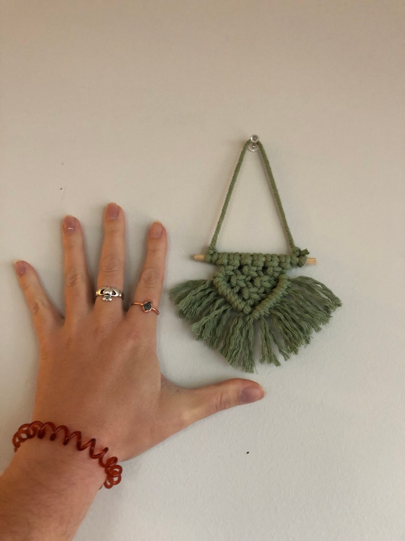 K&ouml;nnte beinhalten: Ein kleiner gr&uuml;ner Makramee-Wandbehang mit Fransen, der an einer wei&szlig;en Wand h&auml;ngt.