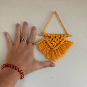 K&ouml;nnte beinhalten: Ein kleiner gelber Makramee-Wandbehang mit Fransen, der an einer wei&szlig;en Wand h&auml;ngt. Der Wandbehang hat die Form eines Dreiecks und ist etwa so gro&szlig; wie eine Hand.