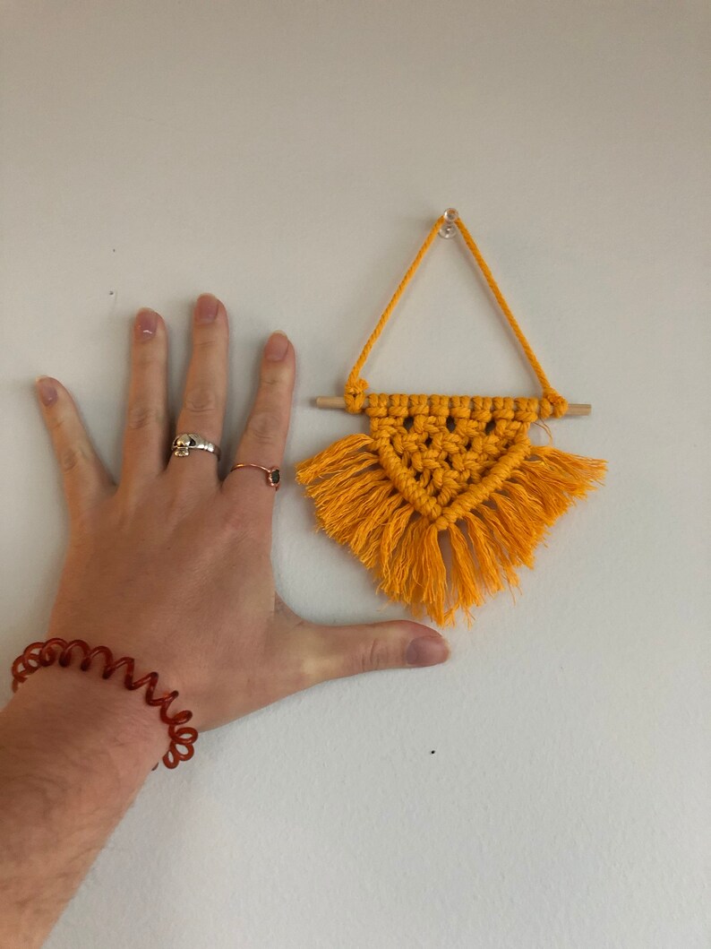 K&ouml;nnte beinhalten: Ein kleiner gelber Makramee-Wandbehang mit Fransen, der an einer wei&szlig;en Wand h&auml;ngt. Der Wandbehang hat die Form eines Dreiecks und ist etwa so gro&szlig; wie eine Hand.
