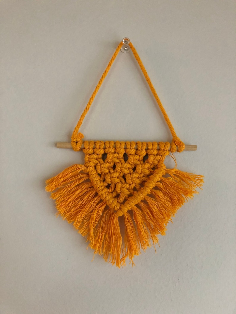 K&ouml;nnte beinhalten: Ein gelber Makramee-Wandbehang mit einem Holzstab und Fransen.