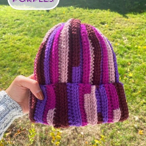 May include: A striped crocheted beanie hat in shades of purple, pink, and brown. The hat has a wide brim and is made with a chunky yarn.