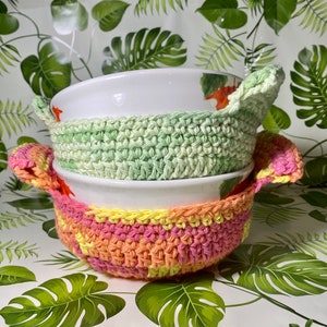 May include: Two white ceramic bowls with a green and pink crocheted bowl cosy. The cosy has a light green top and a pink and orange bottom.