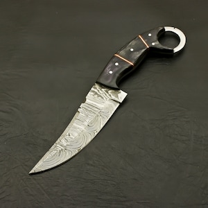 May include: A custom-made knife with a curved blade and a dark handle. The blade features a unique swirling pattern, and the handle has a finger ring. Copper accents and white dots adorn the handle.
