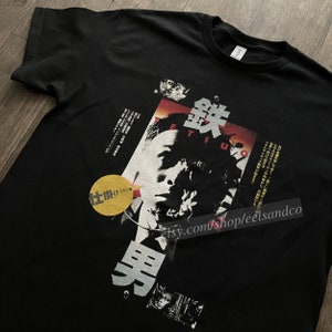 Puede incluir: Camiseta negra con un dise&ntilde;o gr&aacute;fico que incorpora texto japon&eacute;s y un retrato. El dise&ntilde;o incluye la palabra "Tetsuo" y otros caracteres japoneses. Tambi&eacute;n hay un c&iacute;rculo amarillo con texto japon&eacute;s.