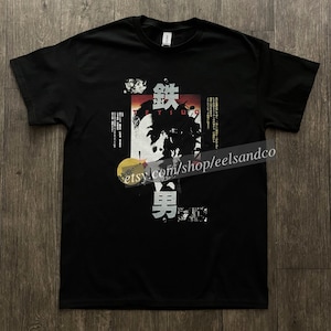 Puede incluir: Camiseta negra con un dise&ntilde;o gr&aacute;fico que incorpora texto japon&eacute;s y un rostro estilizado. El dise&ntilde;o utiliza elementos rojos, blancos y negros. La palabra "TETSUO" es visible.