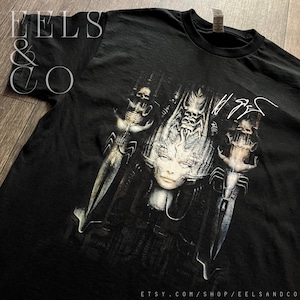 Puede incluir: Camiseta negra con un gráfico surrealista detallado que presenta figuras esqueléticas y una cara central. El diseño es predominantemente en escala de grises con detalles intrincados. La URL de la tienda Etsy es visible en la parte inferior.