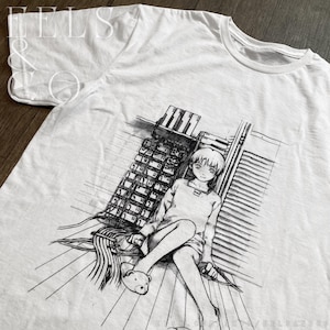 May include: White t-shirt featuring a black and white anime-style illustration. The graphic depicts a seated figure with short hair, wearing a long-sleeved shirt and shorts, with a teddy bear slipper. The background includes a building and a window.