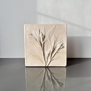 May include: A square, cream-coloured decorative tile with a detailed relief of a floral arrangement. The design includes long leaves and delicate flower buds. The tile is displayed on a reflective surface.