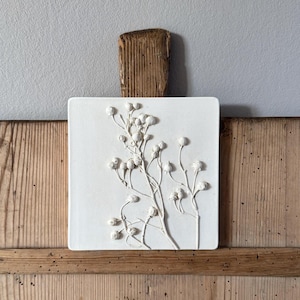 Könnte beinhalten: Quadratische, weiße Keramik-Wandkunst mit einem erhabenen Relief aus zarten, weißen Blütenstängeln und Knospen. Das Kunstwerk ist auf einem Stück verwittertem Holz mit natürlicher Holzmaserung montiert.