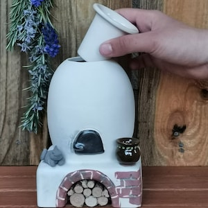 May include: A white ceramic oven with a black door, a small brown pot with green accents, and a grey cat figurine. The oven has a brick-like design and a wood-burning stove effect. The oven is sitting on a wooden surface.