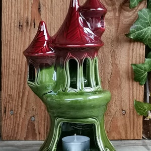 Pode incluir: Um porta-velas de cerâmica em forma de castelo com telhado vermelho e corpo verde. O porta-velas tem uma pequena abertura na parte inferior para uma vela de chá.