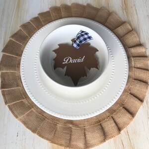 May include: A place setting with a white bowl and plate, a brown leaf-shaped name card with the name "David", and a burlap placemat. The name card is tied with a black and white checkered ribbon. The plate has a beaded edge.