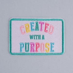 May include: A light pink embroidered patch with a teal border. The text "CREATED WITH A PURPOSE" is embroidered in rainbow colors.