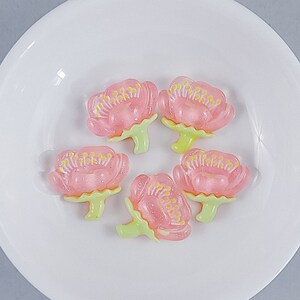 May include: Five pink flower-shaped candies with yellow centers and green stems are arranged on a white plate. The candies have a translucent appearance, and the plate is round.