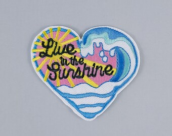 May include: Embroidered iron-on patch in the shape of a heart with a sun and wave design. The patch is pink, yellow, blue, and white. The text on the patch reads "Live in the Sunshine."