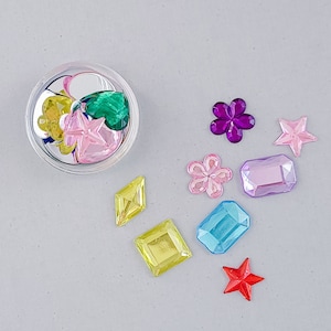 May include: Assortment of colourful plastic gems in various shapes, including stars, flowers, and diamonds. The gems are in a clear plastic container and on a grey surface.