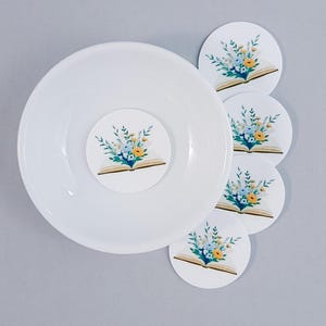 Könnte beinhalten: Eine weiße Keramikschale mit floralem Design im Inneren und vier passenden Untersetzern. Jeder Untersetzer und die Mitte der Schale zeigen ein offenes Buch mit einem Strauß blauer, gelber und grüner Blumen.