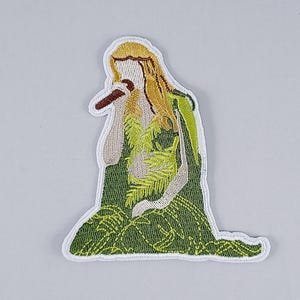 May include: Embroidered patch featuring a woman with long blonde hair wearing a green and yellow dress. She is holding a microphone and singing.