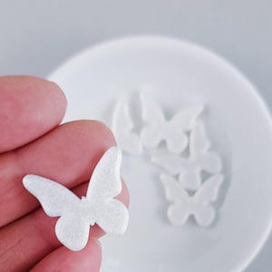 Op de afbeelding: Een kleine, witte, vlindervormige versiering met een subtiele glinsterende textuur. De vlinder heeft afgeronde vleugels en een eenvoudig, symmetrisch silhouet. Verschillende identieke witte vlinderstukjes zijn op de achtergrond zichtbaar.