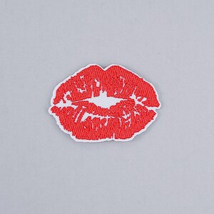 May include: An embroidered patch featuring a vibrant red lip print design. The lips are depicted with a textured, stitched appearance, showcasing a detailed pattern of red thread against a white background, creating a clear, bold graphic shape.