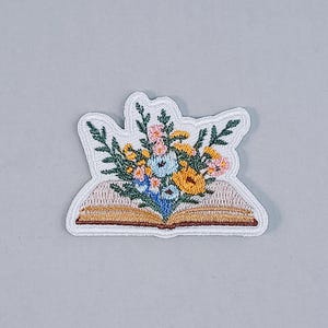 Könnte beinhalten: Gestickter Aufnäher mit einem offenen Buch und einem bunten Blumenstrauß. Die Blumen sind rosa, gelb und blau, mit grünen Blättern. Der Aufnäher hat einen weißen Rand und befindet sich auf einem hellgrauen Hintergrund.
