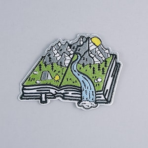 Op de afbeelding: Een geborduurde patch die een open boek voorstelt met een bergketen, een rivier en een kleine camping erin. Het boek is zwart omrand en de bergen zijn grijs. De rivier is blauw en de camping is groen.