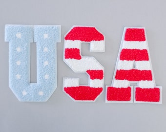 Peut inclure: USA écrit en grandes lettres en feutre. Les lettres sont rouge, blanc et bleu. Le U est bleu avec des étoiles blanches, le S et le A sont rouges avec des rayures blanches.
