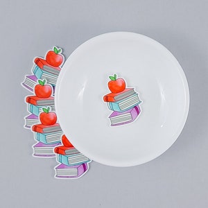 May include: A white plate with a stack of books and a red apple on top. The books are purple, blue, and white. There are several stacks of books and apples around the plate.
