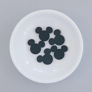 Könnte beinhalten: Eine weiße Keramikschale enthält fünf schwarze, Mickey Mouse-förmige Ausschnitte. Die Schale ist rund mit einem leicht erhöhten Rand, und die Ausschnitte sind gleichmäßig verteilt, vor einem hellgrauen Hintergrund.